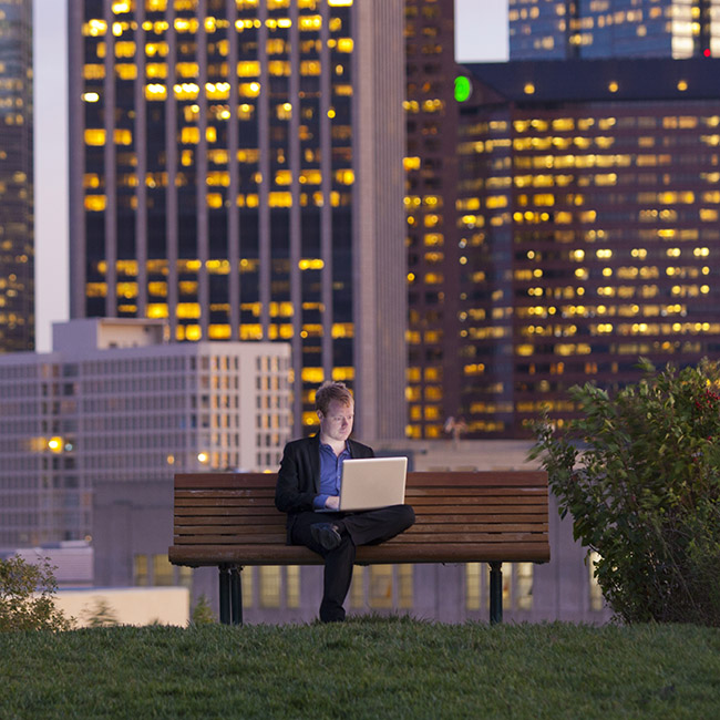 Un homme est assis sur un banc dans un parc au milieu d'une zone urbaine, travaillant sur un ordinateur portable.