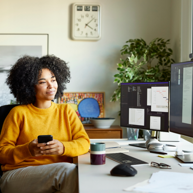 Uma mulher concentrada no trabalho, sentada em uma mesa com um computador e um telefone celular por perto, realizando multitarefas com eficiência.