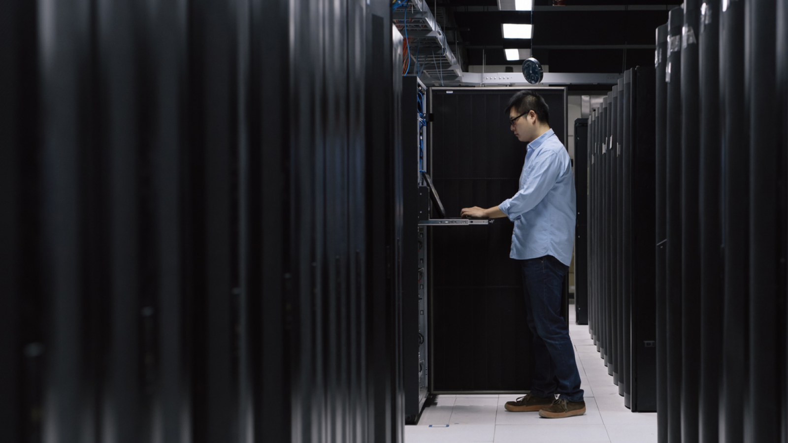 A man stands in a server room, focused on his laptop, managing data and ensuring smooth operations.