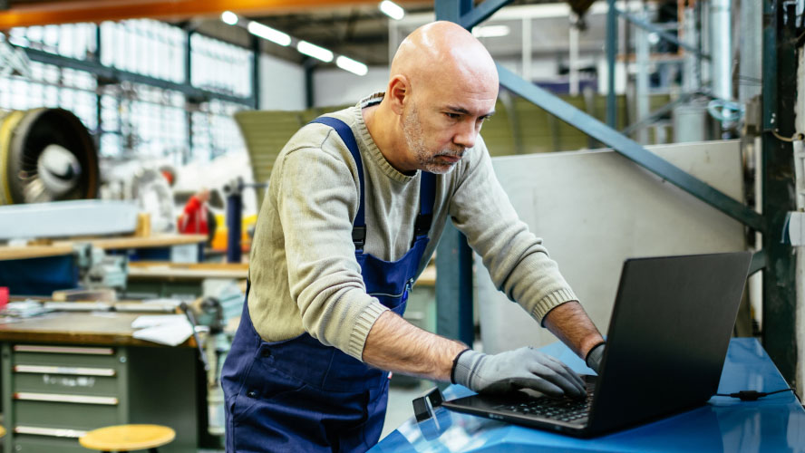 Ein Mann in einer Industrieumgebung arbeitet konzentriert an seinem Laptop und erledigt fleißig Aufgaben.