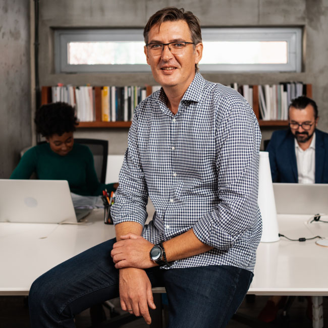 Un uomo con gli occhiali siede a una scrivania con due computer portatili, impegnato nel lavoro, mostrando concentrazione e produttività.