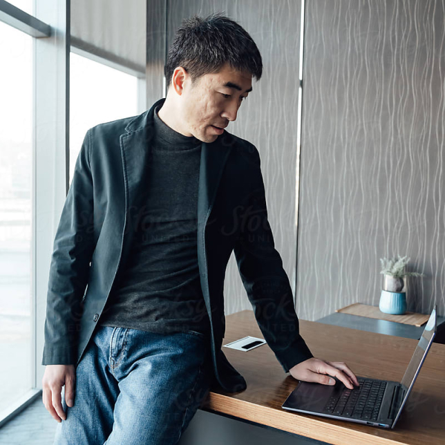 Um homem em traje casual, vestindo jeans e jaqueta, está sentado em uma mesa, concentrado em seu trabalho em um laptop.